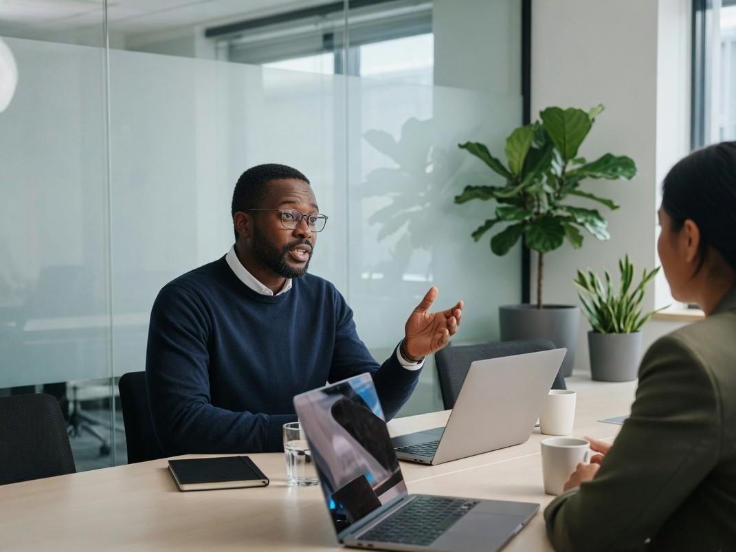 Two colleagues discussing a workplace program at a conference table, laptop screens turned away from the camera